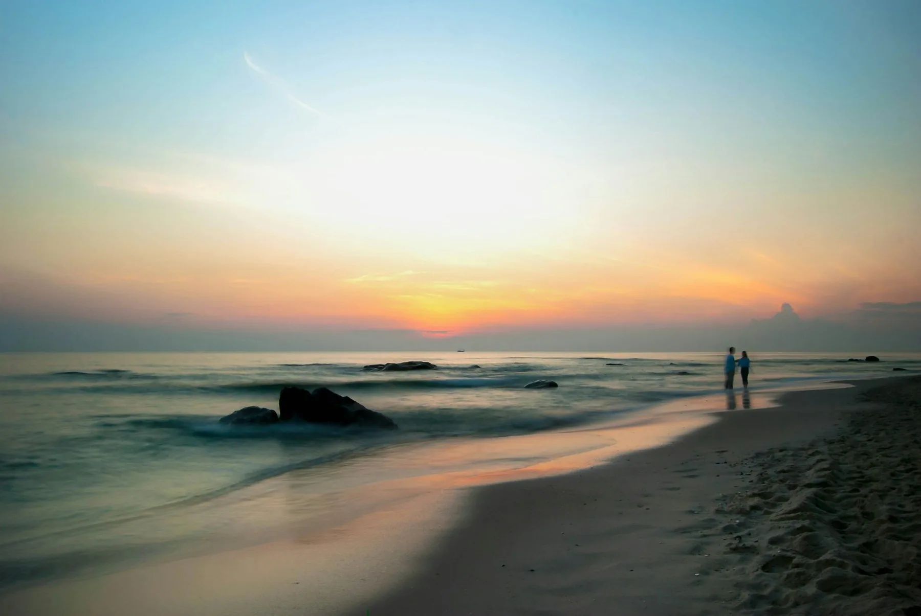 Solnedgang over en stille strand i Hua Hin med steiner i vannkanten og to personer på sanden
